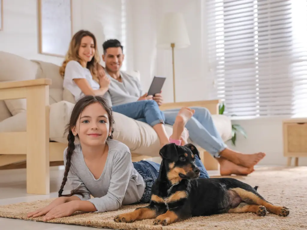 A young girl lies on a rug with her dog, her parents sitting together on a couch in the background. The space is modern, clean and tidy.