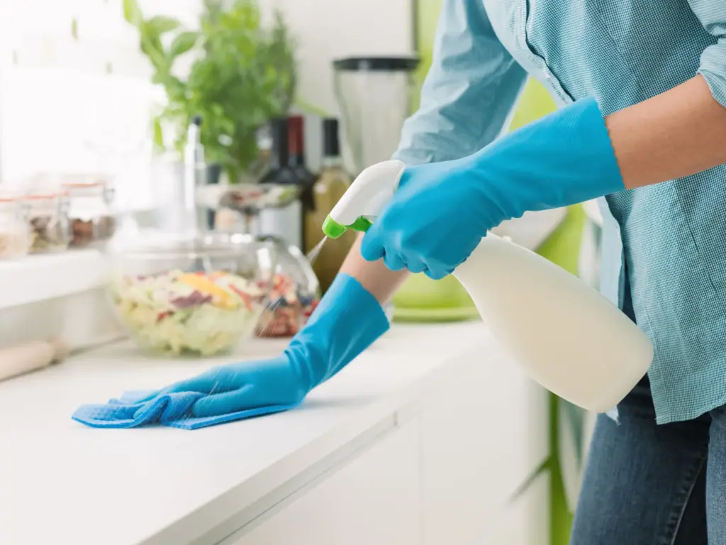 A cleaner wearing blue gloves and holding a spray bottle wipes a kitchen benchtop. Only the hands and arms are visible.