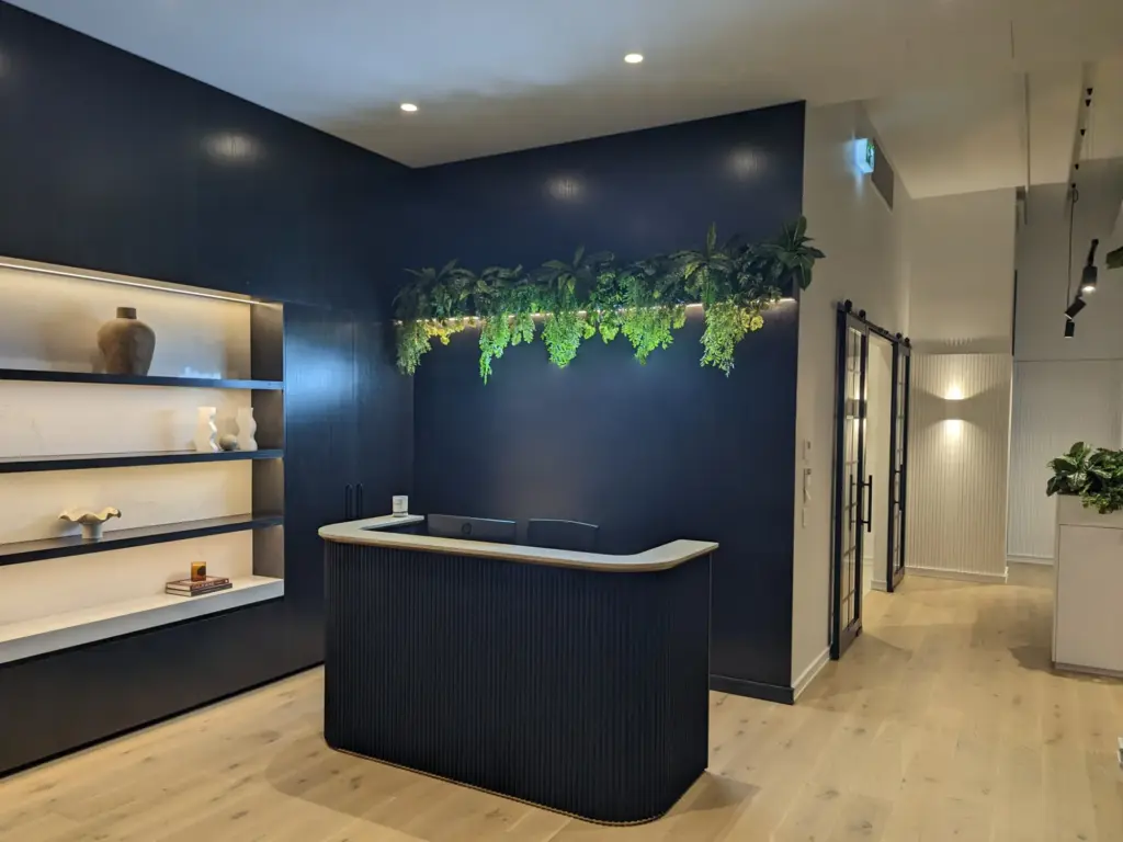 The foyer entrance to a beautifully clean workplace. Dark wood paneled walls and reception desk, and illuminated shelf, light polished wooden floors.