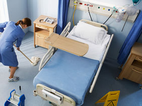 A cleaner mopping the floor under a patient bed in a hospital.